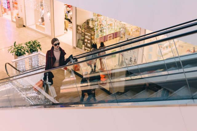 hic woman in sunglasses holds bags up a mall escalator